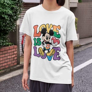 Op de afbeelding: Wit t-shirt met Mickey Mouse die een rood hart vasthoudt. De tekst "LOVE IS LOVE" staat in kleurrijke letters in retrostijl. Het ontwerp bevat regenboogkleuren en een cartoonstijl.