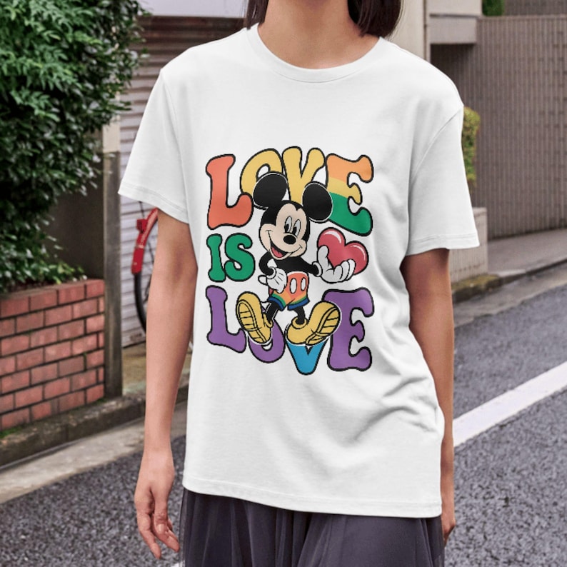 Op de afbeelding: Wit t-shirt met Mickey Mouse die een rood hart vasthoudt. De tekst "LOVE IS LOVE" staat in kleurrijke letters in retrostijl. Het ontwerp bevat regenboogkleuren en een cartoonstijl.