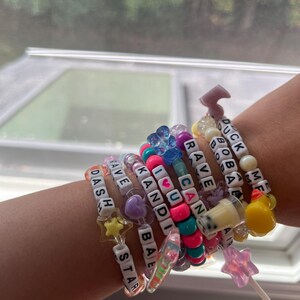 May include: A collection of colorful beaded bracelets with letter beads spelling out phrases like "I Love You", "Can", "Rave", "Dash", "Have", "Kandy", "Bae", and "Star". The bracelets are worn on a person's wrist.