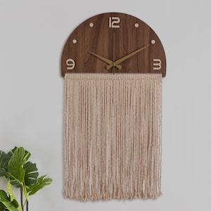 May include: A wooden clock with a half-circle design and a fringe of beige yarn hanging below. The clock face has Roman numerals and gold hands.