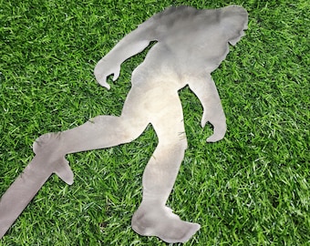 May include: A metal silhouette of a Bigfoot walking, cut out from a sheet of metal. The silhouette is standing on a green artificial grass surface.