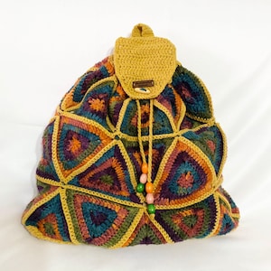 Può includere: Uno zaino all'uncinetto con un motivo geometrico in tonalità di marrone, verde, viola e arancione. Lo zaino ha una chiusura a coulisse e un piccolo lembo con un bottone.