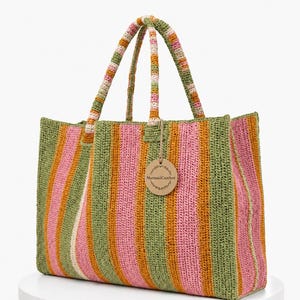 May include: A rectangular crochet tote bag featuring vertical stripes in olive green, bright pink, and mustard yellow. The bag has two matching striped handles and a round brown paper tag attached with string that reads MermaidCrochet.