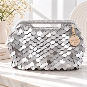 May include: A silver crochet handbag covered in overlapping circular metallic sequins. The bag features a structured top handle and a round wooden tag attached with a light-colored strap, clearly engraved with the text Mermaid Crochet.