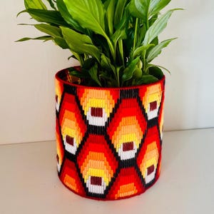 May include: A colourful, hand-stitched plant pot featuring a geometric design in red, orange, yellow, and black. The pot holds a thriving green plant with large leaves. The pot is a decorative container for indoor plants.