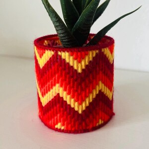 May include: A small, red, orange and yellow crocheted plant pot with a chevron pattern. A small green succulent plant is inside the pot.