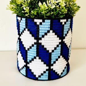 May include: A cylindrical plant pot with a geometric pattern in shades of blue, white, and black. The pot is filled with green artificial plants. The design features diamond shapes, creating a visually appealing texture. Suitable for indoor use.