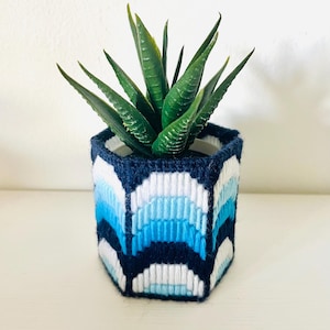 May include: A hexagonal plant pot cover featuring a stitched yarn pattern in shades of navy blue, light blue, and white. The geometric design consists of repeating arched shapes arranged in horizontal bands, housing a green succulent plant.