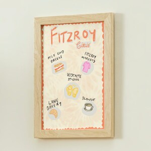 May include: A framed print with an orange background and white text that reads "Fitzroy Girlie". The print features illustrations of various local businesses, including Mile End Bagels, Fitzroy Markets, Upstate Studios, Lune Bakery, and Blonde.