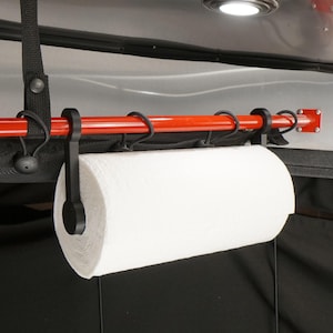 May include: A black paper towel holder with a red metal bar and black hooks. The holder is mounted to a black surface and has a white paper towel roll attached.