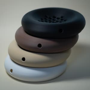 May include: A stack of four ceramic, circular objects in varying colours: white, beige, brown, and black. Each object has a hole on the side. The top object has a perforated surface.