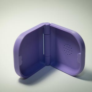 Puede incluir: Una funda morada con bisagras y bordes redondeados. La funda est&aacute; hecha de un material liso y tiene un peque&ntilde;o dise&ntilde;o circular similar a un altavoz en un lado. La funda est&aacute; abierta, revelando el interior.
