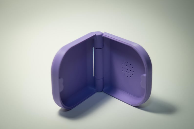 Puede incluir: Una funda morada con bisagras y bordes redondeados. La funda est&aacute; hecha de un material liso y tiene un peque&ntilde;o dise&ntilde;o circular similar a un altavoz en un lado. La funda est&aacute; abierta, revelando el interior.