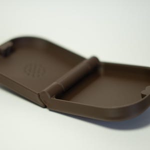 Puede incluir: Un estuche marr&oacute;n, abierto y con bisagras. El estuche es rectangular con esquinas redondeadas y un acabado mate. El interior tiene una superficie lisa. La bisagra es una barra cil&iacute;ndrica. El estuche est&aacute; dise&ntilde;ado para guardar objetos peque&ntilde;os.