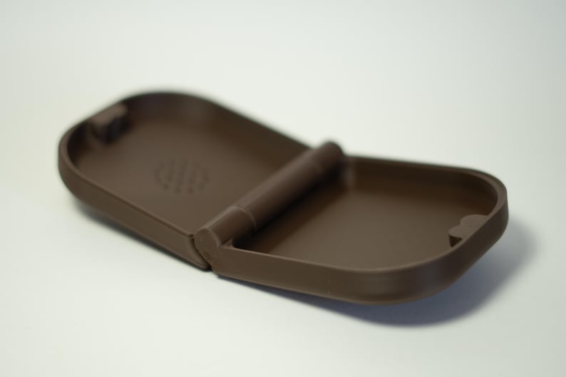 Puede incluir: Un estuche marr&oacute;n, abierto y con bisagras. El estuche es rectangular con esquinas redondeadas y un acabado mate. El interior tiene una superficie lisa. La bisagra es una barra cil&iacute;ndrica. El estuche est&aacute; dise&ntilde;ado para guardar objetos peque&ntilde;os.