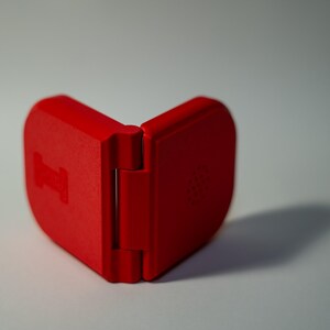 Puede incluir: Un estuche rojo brillante, con bisagras, abierto para revelar su interior. El estuche tiene una forma redondeada y un acabado mate. La bisagra es visible y el estuche proyecta una sombra sobre la superficie blanca.