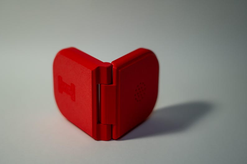 Puede incluir: Un estuche rojo brillante, con bisagras, abierto para revelar su interior. El estuche tiene una forma redondeada y un acabado mate. La bisagra es visible y el estuche proyecta una sombra sobre la superficie blanca.