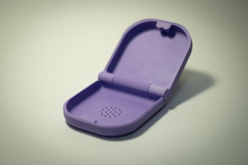 Puede incluir: Estuche lavanda abierto con tapa abisagrada. El estuche tiene forma rectangular redondeada con una serie de peque&ntilde;os agujeros cerca de la parte inferior. El estuche est&aacute; vac&iacute;o y se encuentra sobre una superficie clara.