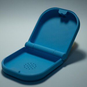 Puede incluir: Un estuche azul claro abierto. El estuche es de material liso y tiene forma rectangular redondeada. El interior tiene un peque&ntilde;o patr&oacute;n circular de agujeros. El estuche est&aacute; abierto, revelando el interior.