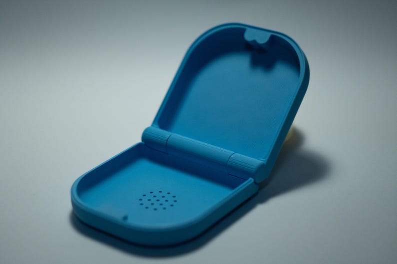 Puede incluir: Un estuche azul claro abierto. El estuche es de material liso y tiene forma rectangular redondeada. El interior tiene un peque&ntilde;o patr&oacute;n circular de agujeros. El estuche est&aacute; abierto, revelando el interior.