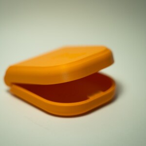 Puede incluir: Un recipiente naranja abierto y con bisagras. El contenedor es rectangular con esquinas redondeadas y una tapa curva. El interior es visible y el contenedor parece estar hecho de pl&aacute;stico.