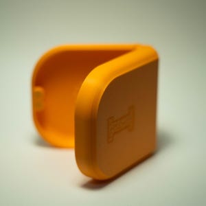 Puede incluir: Carcasa naranja brillante, con bisagras y forma rectangular redondeada. La carcasa est&aacute; abierta, revelando el interior. Un peque&ntilde;o dise&ntilde;o en relieve es visible en la parte delantera. La carcasa est&aacute; hecha de un material liso y s&oacute;lido.