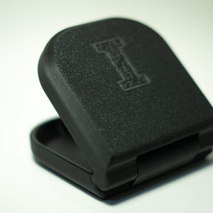 Puede incluir: Un estuche negro con bisagras y superficie texturizada. La parte superior del estuche presenta la letra "I" en relieve. El estuche est&aacute; abierto, revelando el interior. El objeto est&aacute; sobre un fondo blanco.