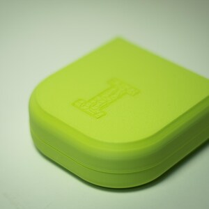 Puede incluir: Un recipiente cuadrado redondeado de color verde lima con un dise&ntilde;o empotrado de la letra "I" en la parte superior. El recipiente tiene un acabado liso y mate y un borde curvo. El objeto est&aacute; sobre un fondo blanco.