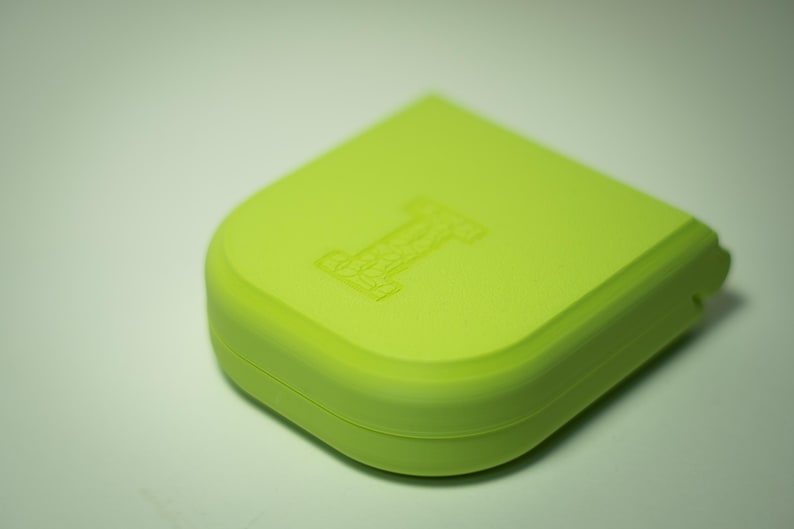 Puede incluir: Un recipiente cuadrado redondeado de color verde lima con un dise&ntilde;o empotrado de la letra "I" en la parte superior. El recipiente tiene un acabado liso y mate y un borde curvo. El objeto est&aacute; sobre un fondo blanco.