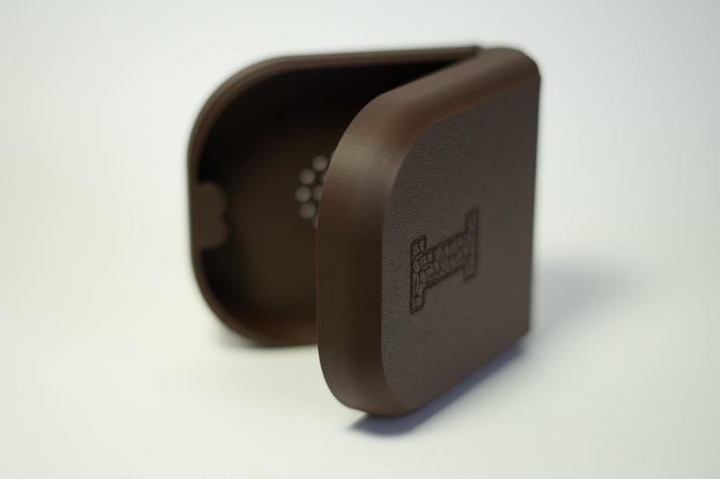 Puede incluir: Una funda marr&oacute;n en forma de concha, abierta para revelar un interior perforado. La funda tiene bordes redondeados y una superficie texturizada. Un logotipo est&aacute; grabado en la parte delantera. Es probable que la funda est&eacute; dise&ntilde;ada para proteger un peque&ntilde;o dispositivo electr&oacute;nico.