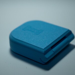 Puede incluir: Peque&ntilde;o objeto rectangular azul claro con superficie texturizada. El objeto tiene una parte superior redondeada y una ranura en un lado. La letra "I" est&aacute; grabada en la superficie superior. El objeto proyecta una sombra.