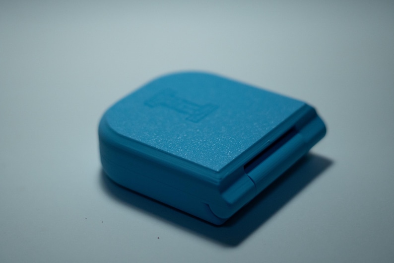 Puede incluir: Peque&ntilde;o objeto rectangular azul claro con superficie texturizada. El objeto tiene una parte superior redondeada y una ranura en un lado. La letra "I" est&aacute; grabada en la superficie superior. El objeto proyecta una sombra.
