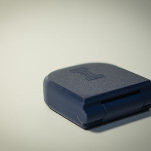 Puede incluir: Un objeto rectangular azul marino con esquinas redondeadas. La superficie superior tiene un aspecto texturizado y un logotipo blanco. El objeto est&aacute; cerrado, con una bisagra visible en el lateral. El art&iacute;culo est&aacute; sobre una superficie blanca.