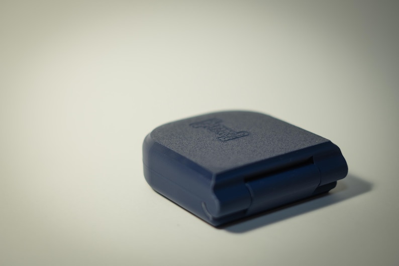 Puede incluir: Un objeto rectangular azul marino con esquinas redondeadas. La superficie superior tiene un aspecto texturizado y un logotipo blanco. El objeto est&aacute; cerrado, con una bisagra visible en el lateral. El art&iacute;culo est&aacute; sobre una superficie blanca.