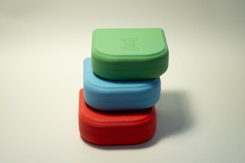 Puede incluir: Tres contenedores cuadrados y coloridos apilados uno encima del otro. El contenedor superior es verde, el del medio es azul y el inferior es rojo. El contenedor verde tiene un logotipo en la tapa.