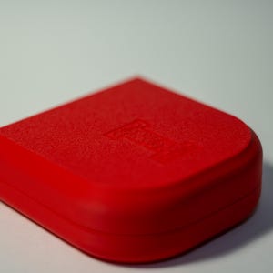 Puede incluir: Un objeto rojo brillante, de forma cuadrada con esquinas redondeadas. La superficie superior tiene una apariencia texturizada, y un dise&ntilde;o sutil es visible. El objeto se encuentra sobre un fondo blanco liso, resaltando su color vibrante y forma.
