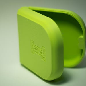 Puede incluir: Un estuche rectangular verde lima con bordes redondeados, parcialmente abierto para revelar un interior verde m&aacute;s claro. El exterior presenta un s&iacute;mbolo de cama en relieve. El estuche est&aacute; hecho de un material mate y liso.