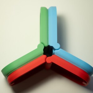 Puede incluir: Objeto abstracto y colorido compuesto por tres piezas entrelazadas. Cada pieza es de un color diferente: verde, rojo y azul. El dise&ntilde;o es geom&eacute;trico y moderno, con bordes redondeados y una disposici&oacute;n sim&eacute;trica.