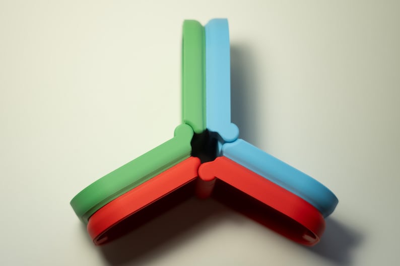 Puede incluir: Objeto abstracto y colorido compuesto por tres piezas entrelazadas. Cada pieza es de un color diferente: verde, rojo y azul. El dise&ntilde;o es geom&eacute;trico y moderno, con bordes redondeados y una disposici&oacute;n sim&eacute;trica.