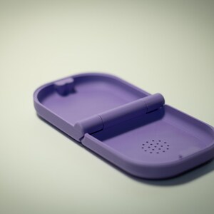 Puede incluir: Un estuche con bisagras de color lavanda, abierto para revelar su interior. El estuche tiene una forma rectangular redondeada con una peque&ntilde;a caracter&iacute;stica circular similar a un altavoz. La bisagra es una barra cil&iacute;ndrica.