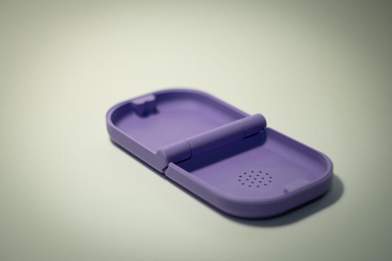 Puede incluir: Un estuche con bisagras de color lavanda, abierto para revelar su interior. El estuche tiene una forma rectangular redondeada con una peque&ntilde;a caracter&iacute;stica circular similar a un altavoz. La bisagra es una barra cil&iacute;ndrica.