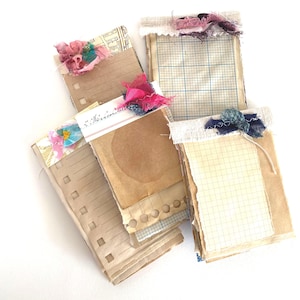 May include: A selection of handmade paper journals with aged, vintage-style pages. Each journal is decorated with colourful fabric and ribbon embellishments. Some pages have grid lines, while others are lined or blank. The journals are stacked, showing their unique textures and designs.