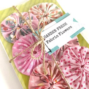 May include: A selection of handmade fabric flowers in various pink hues and floral designs, bound with twine. The flowers are presented on a green and yellow striped base, with a tag reading "GARDEN FRESH Fabric Flowers."