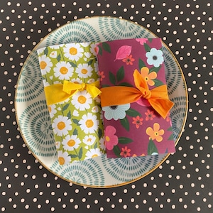 May include: Two floral-patterned notebooks, one with white daisies on green and the other with colorful flowers on a burgundy background, are tied with yellow and orange ribbons. They rest on a decorative plate with a blue and white design, set against a black polka dot background.