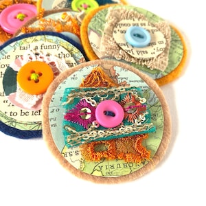 Puede incluir: Una colección de botones de tela hechos a mano con diversas texturas y colores. Cada botón presenta un diseño circular con una impresión de mapa, adornado con retazos de tela coloridos, lentejuelas y botones en tonos rosa, naranja y azul. Algunos botones tienen texto.