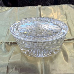 May include: A clear, oval-shaped crystal candy dish with a lid. The dish features a detailed, cut-glass design with a starburst pattern on the lid and geometric shapes on the sides. The dish is sitting on a gold-colored, textured fabric.