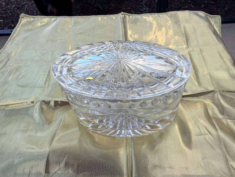 May include: A clear, oval-shaped crystal candy dish with a lid. The dish features a detailed, cut-glass design with a starburst pattern on the lid and geometric shapes on the sides. The dish is sitting on a gold-colored, textured fabric.