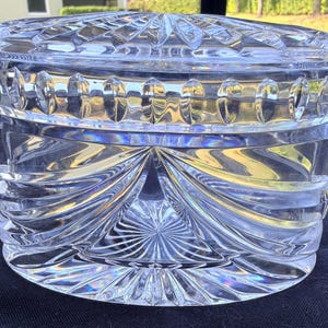 May include: A clear, round, crystal trinket box with a lid. The box features intricate, geometric patterns and a starburst design on the base. The lid has a similar pattern. The box is made of thick, clear glass, reflecting light.