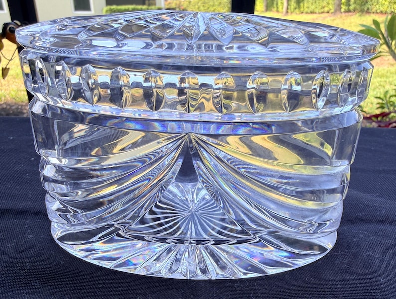 May include: A clear, round, crystal trinket box with a lid. The box features intricate, geometric patterns and a starburst design on the base. The lid has a similar pattern. The box is made of thick, clear glass, reflecting light.