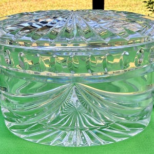 May include: A clear, oval-shaped crystal container with a matching lid. The lid and body feature intricate, geometric patterns, including radial designs and curved lines. The container sits on a green surface, reflecting light and creating a sparkling effect.